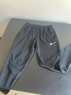 Svarta träningsbyxor från Nike - Säljer ett par svarta träningsbyxor från Nike med vit logga på vänster ben. Byxorna har resår i midjan och är tillverkade i ett mjukt och stretchigt material som passar perfekt för träning eller chill. Snygg och enkel design som funkar till det mesta.