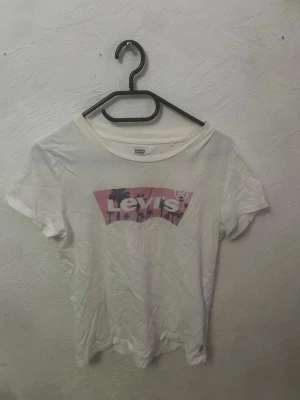 Vit Levi's t-shirt med palmtryck - Säljer en vit t-shirt från Levi's med ett stort tryck framtill i rosa och lila toner, där det står Levi's och motiv av palmer. 