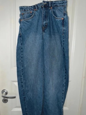 Blåa raka jeans med hög midja - Klassiska blå jeans med rak passform och hög midja. Jeansen har fyra fickor, bälteshällor och knappgylf. Tillverkade i kraftigt denimtyg som ger en tidlös look. Perfekta för dig som gillar vintagevibbar och enkel stil.