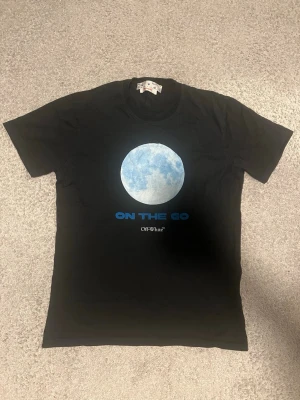 Svart Off-White t-shirt med tryck - Säljer en svart t-shirt från Off-White med ett stort blått cirkeltryck och texten 'ON THE GO' på bröstet. T-shirten har klassisk passform och rund hals. Perfekt för dig som gillar streetwear och vill sticka ut med en unik design.