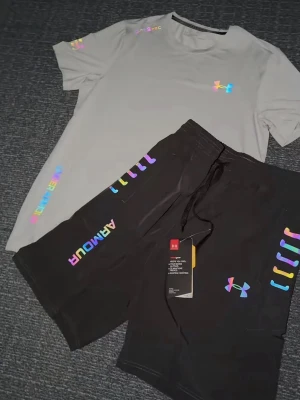 Grå träningsset från Under Armour - Säljer ett par grå träningsset från Under Armour med färgglada loggor och detaljer. Shortsen har snörning i midjan och är tillverkade i ett mjukt, syntetiskt material som passar perfekt för träning eller avslappning. Storlek S, helt ny 