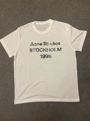 Vit Acne Studios t-shirt Stockholm - Vit t-shirt från Acne Studios med svart tryck på bröstet där det står 'Acne Studios STOCKHOLM 1996'. Klassisk rund hals och korta ärmar. Tillverkad i mjuk bomull för en skön och avslappnad känsla.