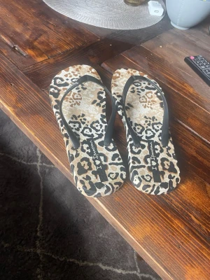 Leopardmönstrade flip-flops från Ipanema - Säljer ett par snygga flip-flops från Ipanema med leopardmönster i beige, brunt och svart. Remmarna är svarta och sandalerna har en klassisk form i mjukt syntetmaterial. Perfekta för sommaren eller stranden! Har blivit använda 1 gång. 