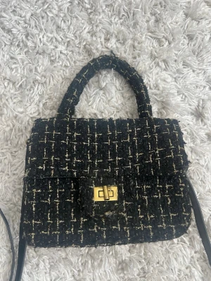 Svart elegant handväska med guldigt lås - Stilren svart handväska i tweed med vita och beige inslag. Väskan har ett klassiskt handtag, axelrem och ett snyggt guldigt vridlås framtill. Perfekt för dig som gillar en chic och trendig look med lite vintagekänsla.