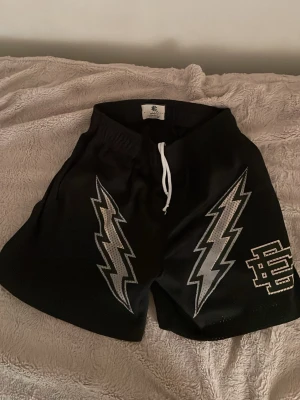 Svarta shorts från Eric Emanuel - Svarta shorts från Eric Emanuel med vita blixtar och logga på ena benet. Shortsen har elastisk midja med vit snörning och är tillverkade i ett lätt, syntetiskt material. Perfekta för en sportig och avslappnad stil.