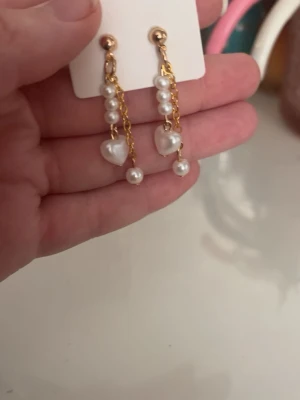 Guldiga örhängen med pärlor och hjärtan - Säljer ett par snygga örhängen med guldfärgade kedjor, vita pärlor och små hjärtformade detaljer. Örhängena har en elegant och trendig look med hängande design och passar perfekt för att lyfta din stil.