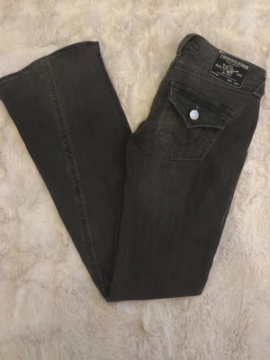 True Religion jeans i modellen ”joey” i färgen grå storlek W25 - Snygga svarta jeans från True Religion med bootcut passform. Jeansen har en klassisk femficksdesign och detaljerad ficka bak med lock och knapp. Tillverkade i mjukt denimtyg som ger en skön känsla och cool look.