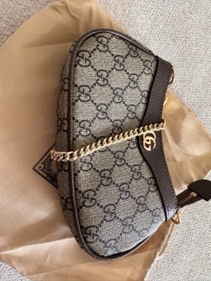 Gucci GG canvas axelremsväska - Snygg handväska från Gucci i beige och brunt GG-mönstrat canvasmaterial med mörkbruna läderdetaljer. Väskan har en guldfärgad kedjerem och GG-logga framtill. Perfekt storlek för det viktigaste och ger en lyxig touch till din outfit.