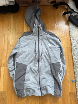 Grå hoodie från The North Face - Snygg grå hoodie från The North Face med dragkedja framtill och kontrasterande mörkgrå paneler på axlar och fickor. Stor huva med dragsko och två sidofickor. Perfekt för chill eller streetwear. Logga på bröstet.