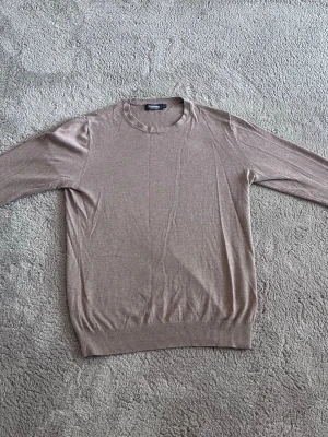 Beige stickad tröja Dressmann - Snygg beige stickad tröja från Dressmann i mjuk bomull. Tröjan har rund halsringning, långa ärmar och ribbade muddar vid ärmslut och nederkant. Perfekt för lager på lager och enkel att matcha med olika outfits.
