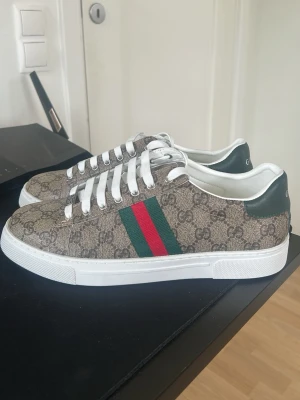 Gucci skor ( HELT OANVÄNDA) - Säljer ett par snygga Gucci sneakers i beige canvas med klassiskt GG-monogram, gröna och röda ränder på sidan samt grönt läder vid hälen där det står GUCCI. Skorna har vita snören och vit sula. Perfekta för dig som vill ha en ikonisk och trendig look.