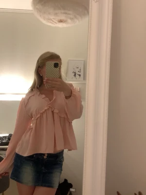 Ljusrosa blus med volangdetaljer - Supersöt ljusrosa blus med långa ärmar och volangdetaljer längs bröstet och halsringningen. Blusen har en lös och flowy passform och är tillverkad i ett tunt, mjukt tyg som känns luftigt. Perfekt för en romantisk och feminin stil. Står ingen storlek men passar mig som har XS. 