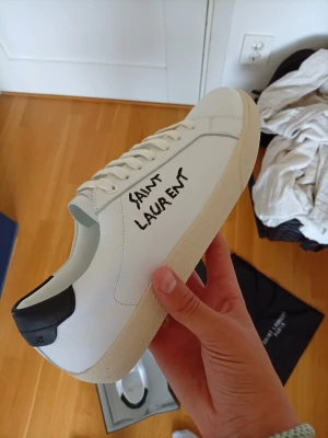 Saint Laurent vita sneakers - Stilrena vita sneakers från Saint Laurent med svart detalj på hälen och handskriven logga på sidan. Skorna har en klassisk låg profil, vita snören och en beige sula. Tillverkade i slätt läder för en clean och modern look.