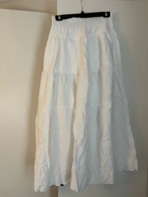 Vit långkjol med broderad volangkant - Säljer en vit långkjol i bomull med bred resår i midjan och flera volanger. Kjolen har broderad spetsdetalj längst ner och är perfekt för varma dagar. Luftig och flowy modell som ger en chill vibe. Kjolen är egensydd.