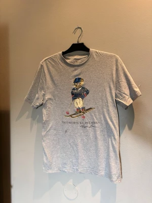 Grå Polo Bear t-shirt från Ralph Lauren - Säljer en grå t-shirt från Ralph Lauren med det ikoniska Polo Bear-trycket framtill där björnen åker skidor i vinteroutfit. T-shirten har rund hals och korta ärmar. Perfekt för dig som gillar klassiska och lekfulla prints.