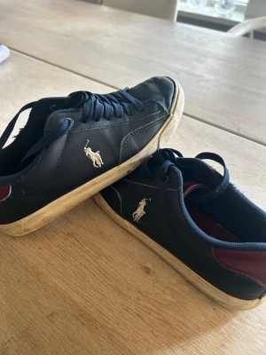 Svarta sneakers med vit logga - Snygga svarta sneakers i läder med mörkblå snören och vit Polo Ralph Lauren-logga på sidan. Skorna har en vit sula och en vinröd detalj vid hälen. Perfekta för en stilren och avslappnad look.