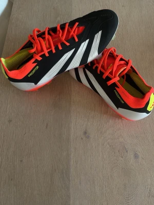 Adidas Predator fotbollsskor svart/vit - Säljer ett par Adidas Predator elite fotbollsskor i svart och vitt med röda detaljer och snörning. Skorna har klassiska tre ränder på sidan, låg profil och dobbar i plast för bra grepp på planen. Snygg design med neonfärgade inslag och texten Predator på sidan och sulan.