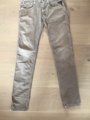 Gråa Replay Anbass Jeans - Beiga slim fit jeans med fem fickor och bälteshällor. Storlek 28/32. Jeansen har raka ben och är gjorda i ett mjukt bomullsmaterial med lite stretch. Snygga sömdetaljer på bakfickorna och klassisk design som funkar till många stilar.