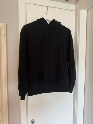 Svart basic hoodie med huva - En enkel svart hoodie med huva och dragsnören. Hoodien har en stor magficka framtill och ribbade muddar vid ärmar och nederkant. 