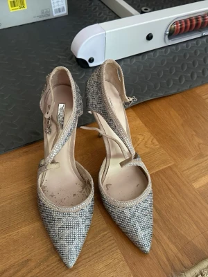 Glittriga pumps med T-rem från Guess - Snygga pumps från Guess med spetsig tå och glittrigt tyg i silver och beige. Skorna har en elegant T-rem och smal klack, samt spänne vid ankeln. Lite skadade inifrån, inget som skadar utseendet eller fötterna. Nypris är 1650kr