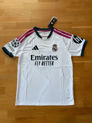 Real Madrid matchtröja Bellingham - Säljer en vit Real Madrid matchtröja med Bellingham och nummer 5 på ryggen. Tröjan har rosa axelränder, mörkgröna detaljer vid ärmslut och hals, samt klubbmärke och sponsortryck framtill. Tillverkad i lätt och ventilerande material, perfekt för fotboll.