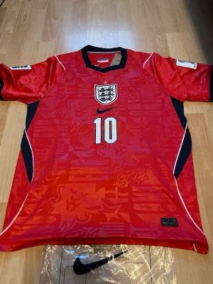 England Bellingham matchtröja röd - Säljer en röd England fotbollströja från Nike med nummer 10 och Bellingham på ryggen. Tröjan har vita och svarta detaljer, broderat landslagsemblem och är tillverkad i ett lätt, ventilerande material. Perfekt för match eller träning.