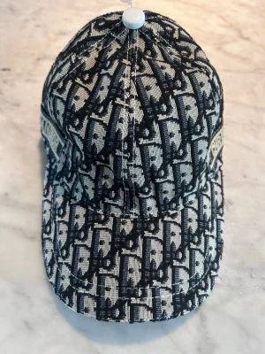 Mönstrad keps  - Snygg keps från  med ikoniskt svart och beige mönster över hela kepsen. Klassisk böjd skärm och broderad logga på sidan. Tillverkad i ett robust tygmaterial som ger en exklusiv känsla. Perfekt för dig som vill sticka ut med en lyxig accessoar.