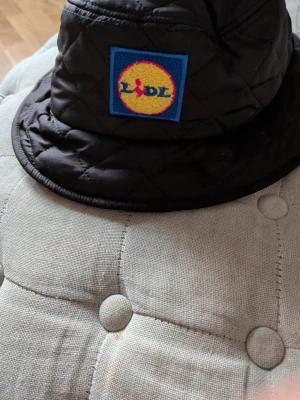 Svart quiltad bucket hat med Lidl-logga - Cool svart quiltad bucket hat med broderad Lidl-logga framtill. Hatten har ett klassiskt bucket hat-snitt och är tillverkad i vadderat material som ger en trendig och sportig look. Perfekt accessoar för att sticka ut i mängden. Väldigt Sällsynt säljs inte längre någonstans!