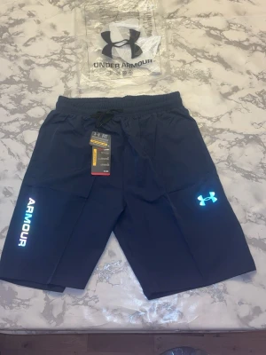 Svarta träningsshorts från Under Armour - Snygga svarta träningsshorts från Under Armour med reflexdetaljer och logga på ena benet. Shortsen har elastisk midja med snörning och är tillverkade i ett lätt och snabbtorkande material. Perfekta för gymmet eller löprundan.