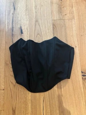 Svart klassisk bustierkorsett - Säljer en svart korsett med klassisk bustierform och markerade sömmar för en snygg siluett. Korsetten är axelbandslös och har en lätt glansig finish. Perfekt att styla till olika looks. Materialet känns som polyester eller liknande.