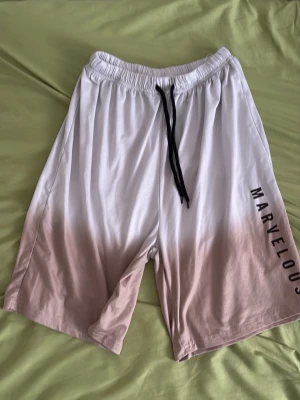 Vita shorts med snörning och tryck - Säljer ett par vita shorts med svart snörning i midjan och texten 'MARVELOUS' tryckt längs ena benet. Shortsen har en relaxed passform och är gjorda i ett mjukt material som passar perfekt till sommaren.