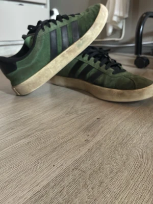Gröna Adidas sneakers i mocka - Snygga gröna Adidas sneakers i mocka med klassiska svarta ränder på sidorna, har små defekter på insidan vid hälen på båda skor men det går lätt att fixa det är bara att lägga i en ny sula. Annars inga stora tecken på användning 
