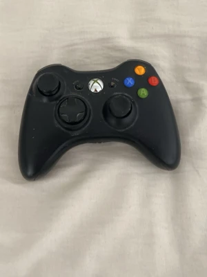 Svart Xbox 360 handkontroll - Säljer en svart Xbox 360 handkontroll med klassisk layout och färgade knappar (grön, röd, blå, gul). Kontrollen har två analoga spakar, styrkors och bekvämt grepp. Perfekt för gaming på konsol eller PC.