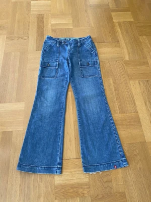 Vintage bootcut cargo jeans - Så fina lågmidjade, utsvängda jeans med cargo detaljer. 99% bomull 1%elastan så de har pyttelite stretch som gör att de sitter snyggt, men fortfarande tjockt och nice jeans material. Perfekt skick förutom liten slitning i hälen på ena benet.  Midja: ca. 39 cm Innerbenslängd: 82 cm Ytterben: 105 cm Benöppning: 27 cm
