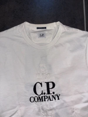 Vit t-shirt från C.P. Company - Vit t-shirt från C.P. Company med broderad logga och diskret motiv framtill. Klassisk rund hals och korta ärmar. Tillverkad i mjuk bomull som känns skön mot huden. Perfekt för dig som gillar stilrena och trendiga plagg.