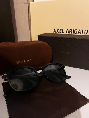 Svarta solglasögon från Tom Ford - Snygga svarta solglasögon från Tom Ford med rundad form och klassisk design. Bågarna är i blank svart plast och glasen är mörka. Kommer med originalfodral och putsduk med Tom Ford-logga. Perfekta för dig som vill ha en stilren accessoar.