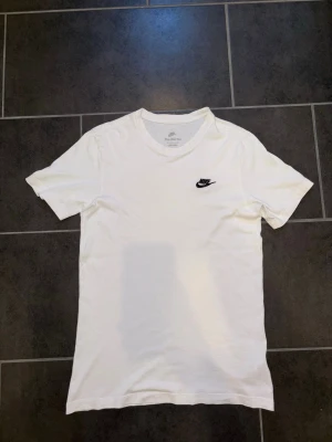 Vit t-shirt från Nike - Klassisk vit t-shirt från Nike med rund halsringning och svart Nike-logga broderad på bröstet. Tillverkad i mjukt bomullsmaterial som känns skönt mot huden. Perfekt basplagg för en avslappnad och sportig stil.