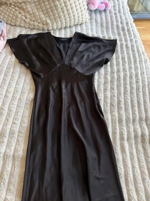 Svart långklänning med v-ringning - Elegant svart långklänning med v-ringad hals och vida, korta ärmar. Klänningen har en smickrande passform och är tillverkad i ett glansigt, mjukt material som ger en lyxig känsla. Perfekt för dig som vill ha en stilren och tidlös look.