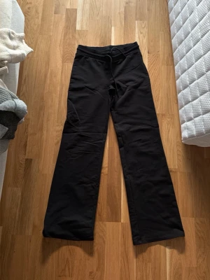 Svarta utsvängda mjukisbyxor - Svarta mjukisbyxor med utsvängda ben och dragsko i midjan. Byxorna har en enkel och stilren design, perfekta för en avslappnad look. Tillverkade i mjukt och bekvämt material som passar för chill dagar.