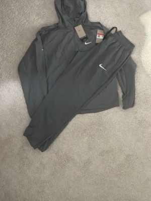 Nike Miller  - Säljer ett mörkgrått loungewear-set från Nike med matchande hoodie och byxor. Setet har en enkel design med Nike-logga i vitt på både tröjan och byxorna. Perfekt för chill dagar hemma. Materialet känns mjukt och bekvämt.