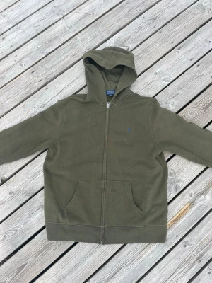 Olivgrön zip Ralph Lauren hoodie - Snygg olivgrön hoodie från Polo Ralph Lauren med dragkedja framtill och klassisk huva. Hoodien har kängurufickor och en liten broderad logga på bröstet. Perfekt för en avslappnad och stilren look.