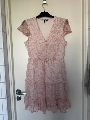 Vero Moda vit och rosa blommig klänning, storlek M - Söt ljusrosa kortklänning med små prickiga mönster, v-ringad hals och knappar framtill. Klänningen har volangdetaljer nertill och korta ärmar. Perfekt för sommarens varma dagar.