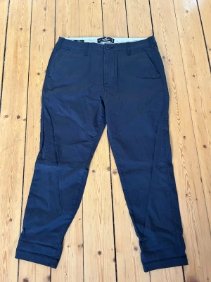 Mörkblå chinos från Hollister - Snygga mörkblå chinos från Hollister med taperad passform och klassiska fickor fram och bak. Tillverkade i ett mjukt och flexibelt material som ger skön komfort hela dagen. Perfekta för en avslappnad men stilren look.