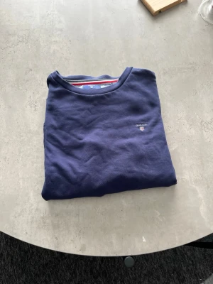 Blå sweatshirt från Gant - Mörkblå sweatshirt från Gant med rund halsringning och diskret logga broderad på bröstet. Tröjan har klassisk passform och ribbade muddar vid ärmslut och nederkant. Perfekt för en avslappnad och stilren look.
