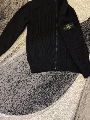 Svart ziptröja från Stone Island - Säljer en svart ribbstickad ziptröja från Stone Island med ikonisk patch på ärmen. Tröjan har hög krage, hel dragkedja framtill och lång ärm. Perfekt för dig som gillar streetwear och vill ha en stilren look.