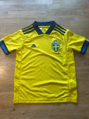 Svensk landslagströja Adidas gul - Äkta svensk landslagströja från Adidas med blå detaljer och tre ränder på axlarna. Tröjan är från säsongen 2021/2022 i barnstorleken 140