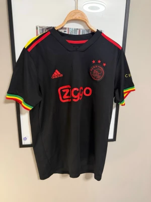 Ajax svart matchtröja Adidas - Snygg svart Ajax matchtröja från Adidas med röda detaljer och Ziggo-logga på bröstet. Tröjan har färgglada ränder i rött, gult och grönt på ärmsluten och axlarna. Ajax-emblemet och Adidas-loggan är broderade i rött. Perfekt för fotbollsfans!