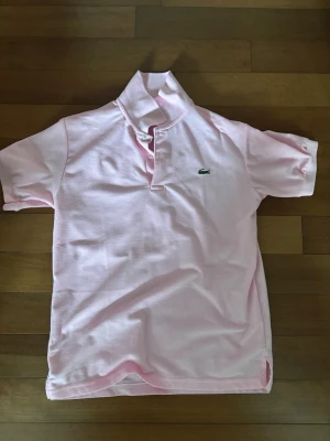 Ljusrosa pikétröja från Lacoste - Säljer en ljusrosa pikétröja från Lacoste med klassisk krage och korta ärmar. Tröjan har den ikoniska gröna krokodilloggan broderad på bröstet och knappar vid halsen. Perfekt för en clean och sportig look.