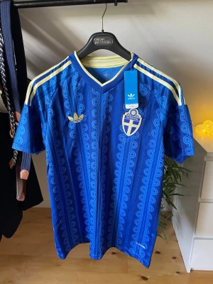 Blå Sverige fotbollströja Adidas - Säljer en blå fotbollströja från Adidas med Sveriges landslagsemblem på bröstet och gula detaljer. Tröjan har ett mönstrat tyg, korta ärmar och klassiska Adidas-ränder på axlarna. Perfekt för dig som vill stötta Sverige med stil. Helt ny, storlek L.