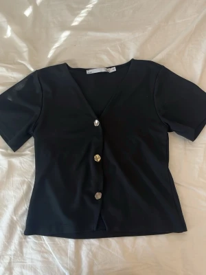 Svart kortärmad topp med knappar - Stilren svart topp med v-ringning och tre dekorativa knappar framtill. Kortärmad modell med en något figurnära passform. Perfekt att matcha med jeans eller kjol för en trendig look.
